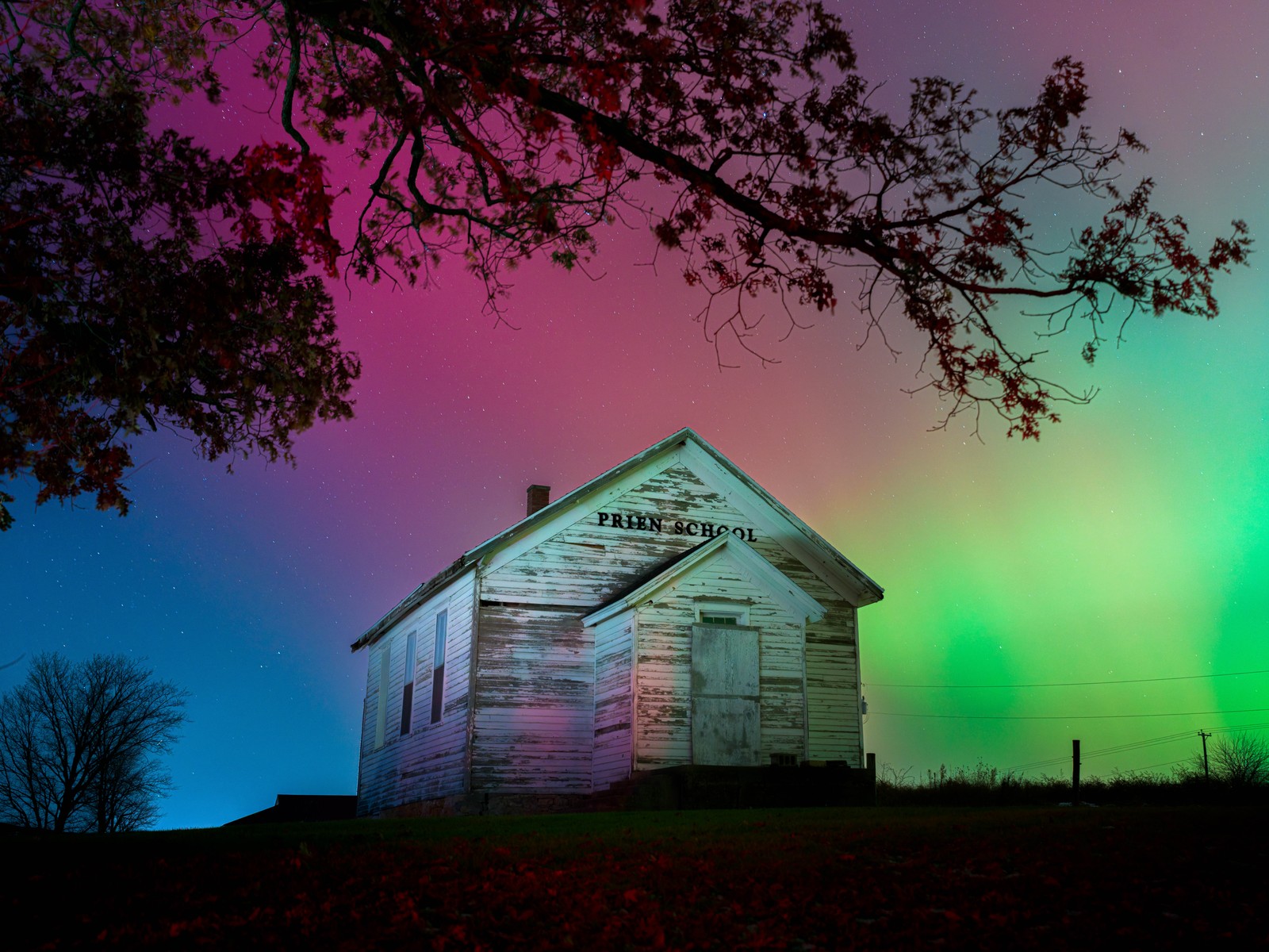 The night sky above a boarded-up wooden school house glows red and green.