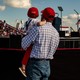 An adult man holds a child at a Trump rally. Both are wearing red hats.