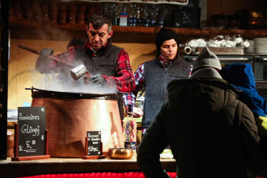 People wait at a market stall, where a person serves hot red wine from a large pot.