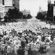 A large crowd of people wearing white gowns and white, pointed hats approaches a fence, against which stand other people in regular clothes.