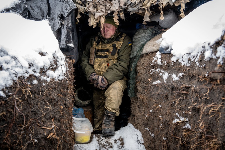 A soldier stands in a narrow trench, beneath a covering, surrounded by snow.