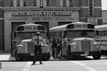 In a black-and-white photo from 1975, black students board school buses lined up in front of South Boston High School. A police office stands in front of one of the busses. 