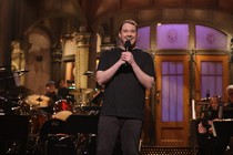 Shane Gillis holding a microphone and smiling onstage during "SNL"