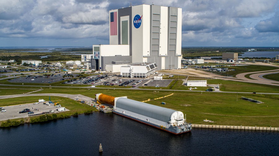 A large section of a rocket is removed from a barge near a very large NASA building used for vehicle assembly.