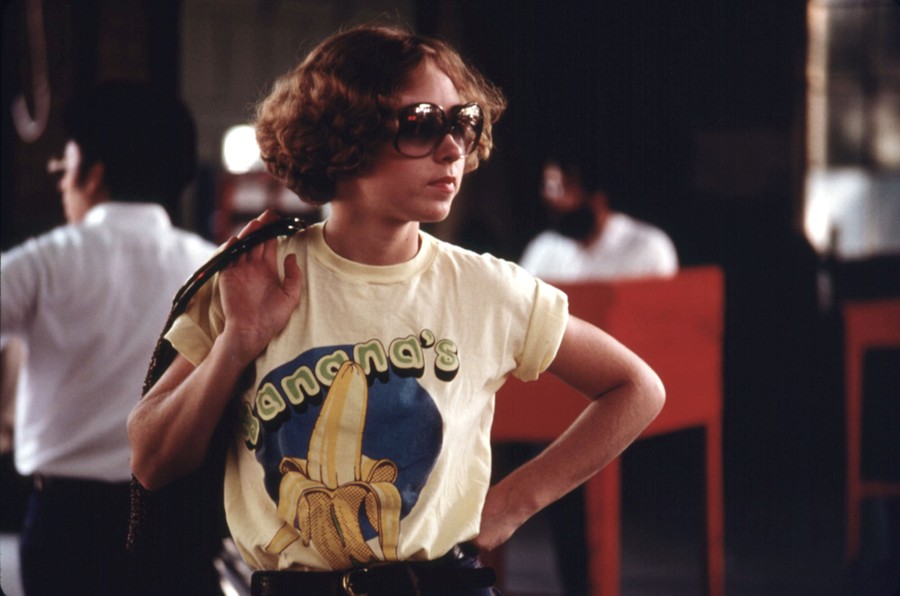 A young woman stands, waiting, wearing large glasses and a t-shirt with an image of a peeled banana on it, and the text "Banana's."