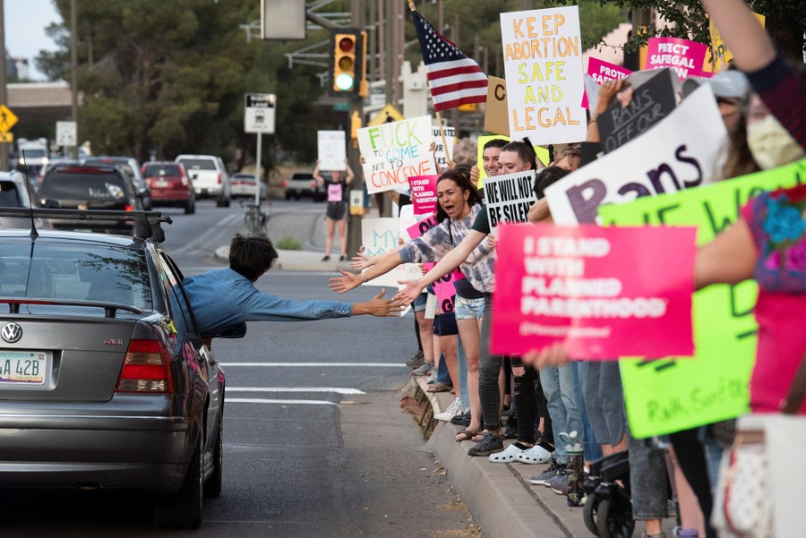 A person leans out the passenger window of a car stretching out their hand to give high fives to a line of protesters standing on a sidewalk.