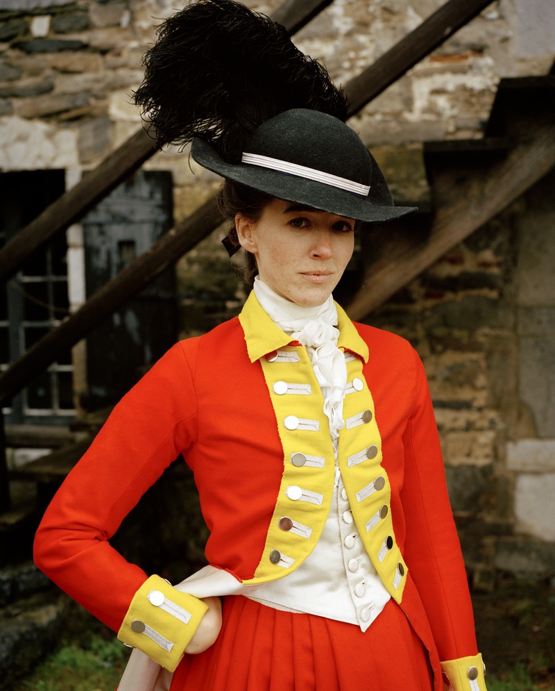 photo of a female reenactor as a British soldier wearing a large plumed black hat, orange-red coat and trousers with yellow lapels and cuffs, standing by wooden stairs with with one hand on hip, inside the grounds of Fort Ticonderoga