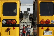 Preschool students play behind a fence. They are framed between two yellow school buses. 