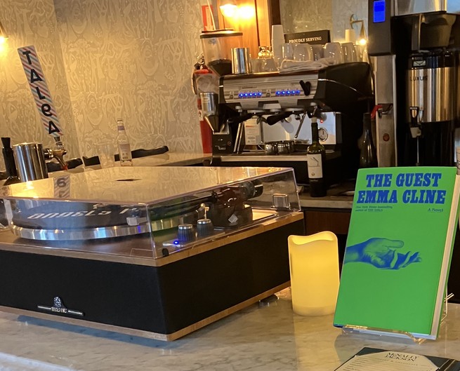 Copy of Emma Cline's The Guest, sitting next to a candle and a record player on the bar at McNally Jackson South Street Seaport location.