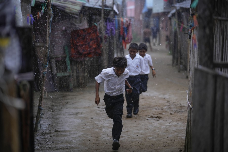 Three boys in school uniforms run down a dirt path between houses during a rainstorm.