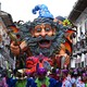 Costumed people parade in front of a large colorful float in the shape of a wizard.