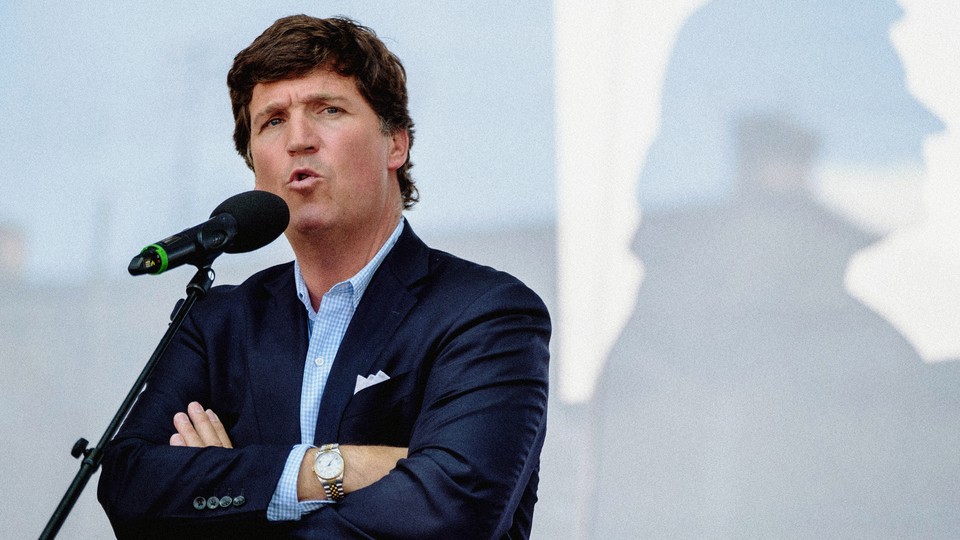 A photo of Tucker Carlson speaking at a microphone with his arms crossed
