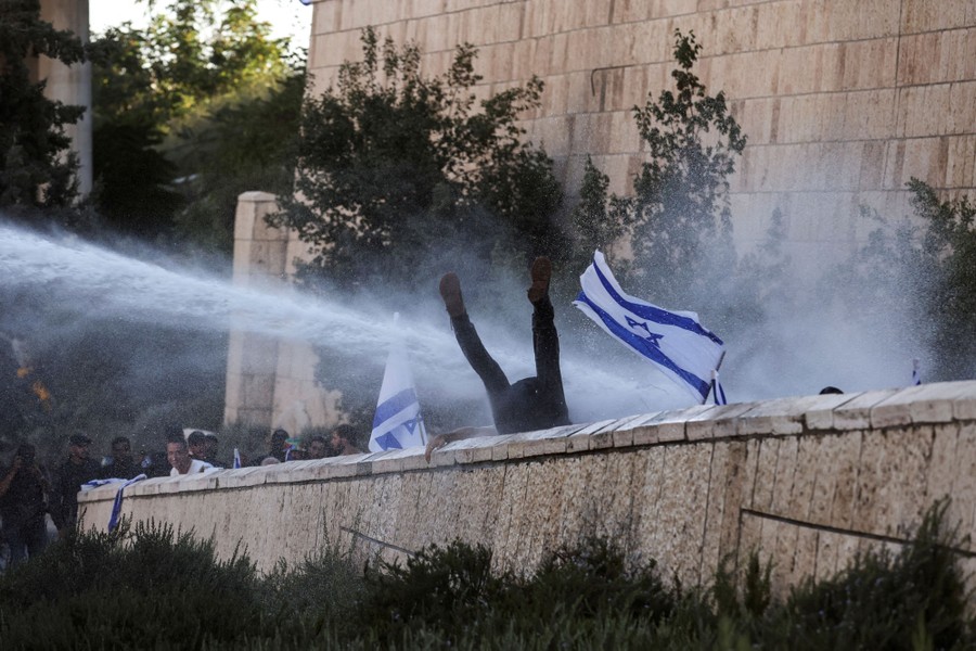 A person who had been sitting on a wall is seen tumbling backward, their legs in the air, after being hit by a water cannon.