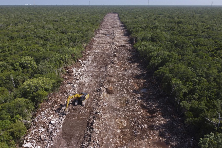 An aerial view of a piece of construction equipment standing in a lane of rocks and dirt dug through a broad surrounding forest.