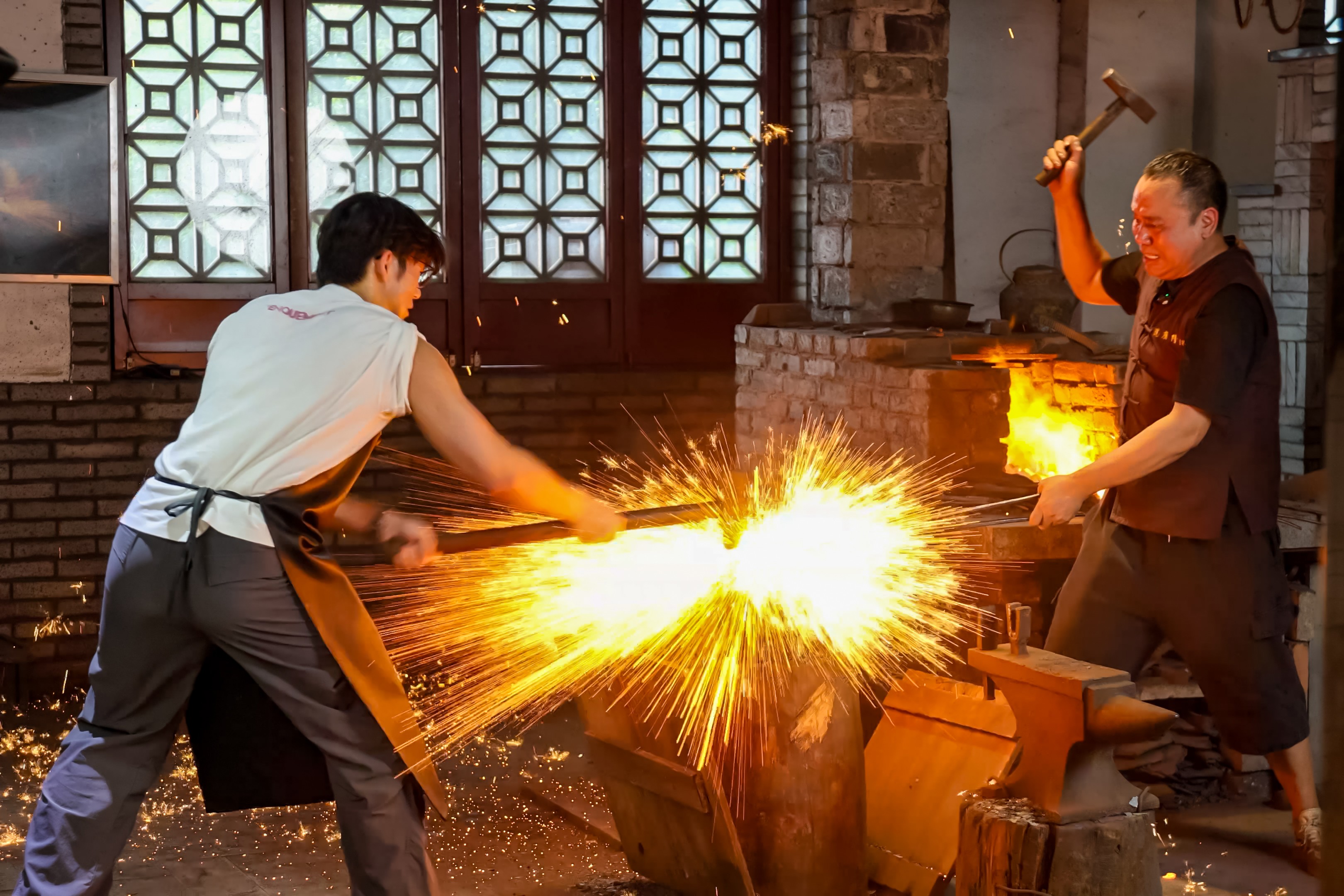 Two workers wield hammers at a sword-making forge, with one striking hot metal and sending up a shower of sparks.