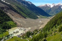 An aerial view of an Alpine valley after a massive landslide, with a cluster of houses being flooded on the uphill side.