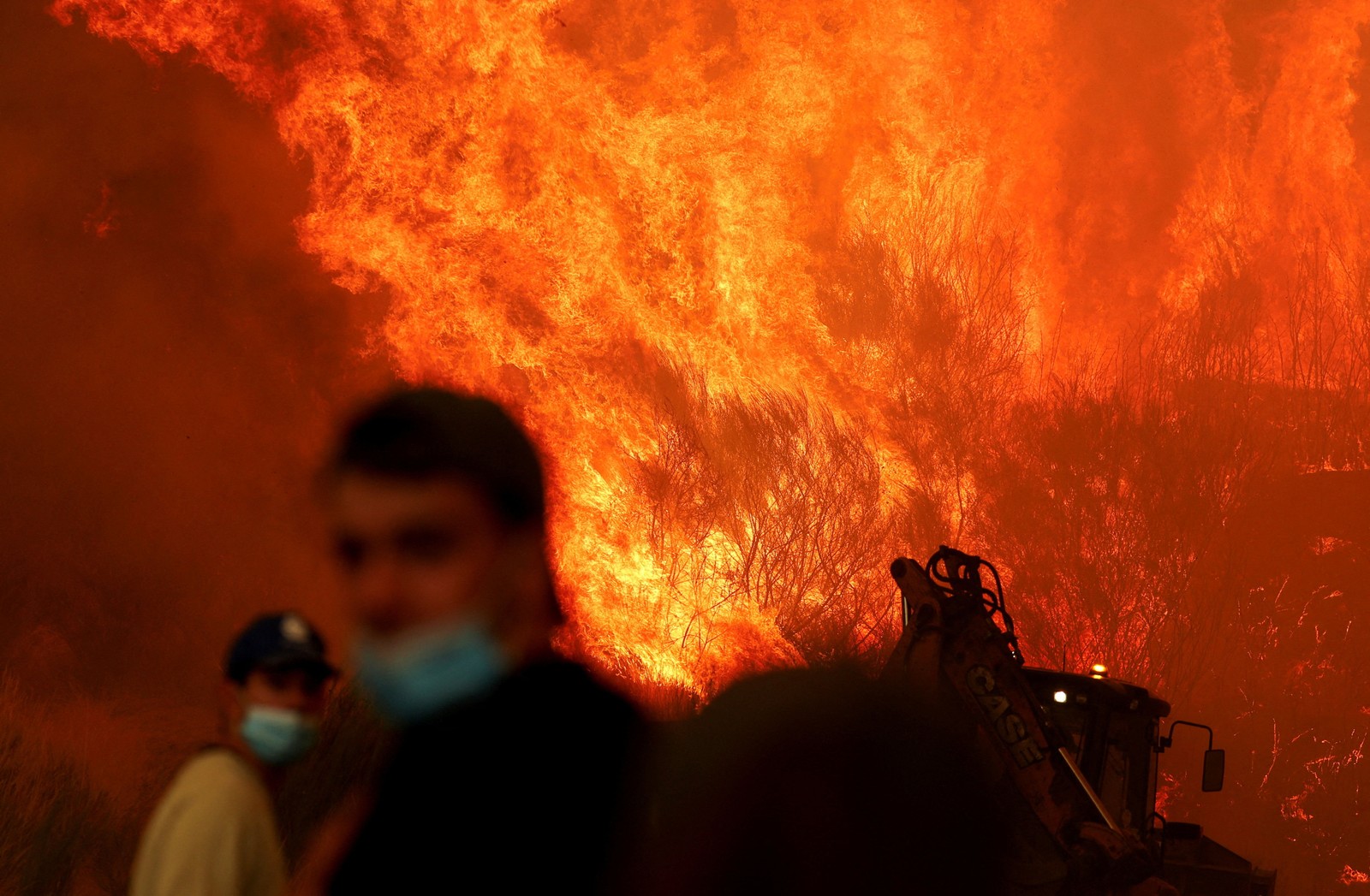 Two people stand in front of a large oncoming wildfire.