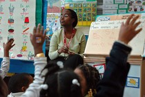 A teacher sits in front of her class. Students raise their hands.