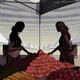 Two women with long hair and large bags, in shadow, reach for tomatoes under a farmers'-market tent