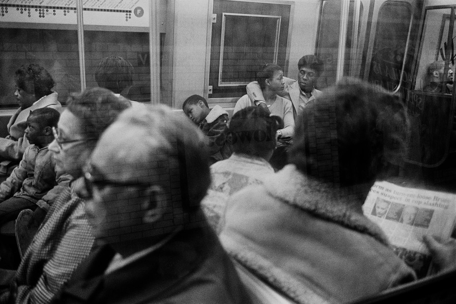 passengers on a subway car seen through the window. A couple with the man's arm around the woman look at each other.