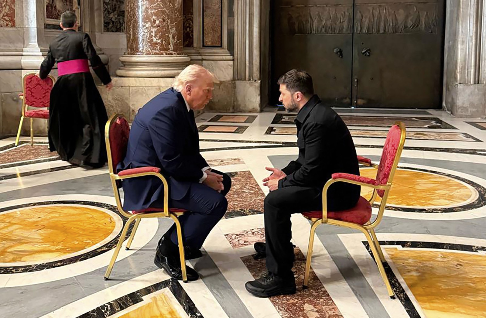 Ukraine's President Volodymyr Zelenskyy and President Donald Trump sit on chairs close together and speak, inside a large room in the Vatican.