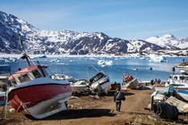 A man walks among boats with snow-covered mountains in the distance.