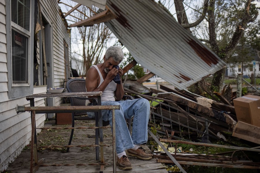 A man sits on his porch and cries with debris from his damaged house visible in the background.