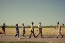 A film still in which a man on the right leads a tour group of five people to his left across a field with barbed wire in the background.