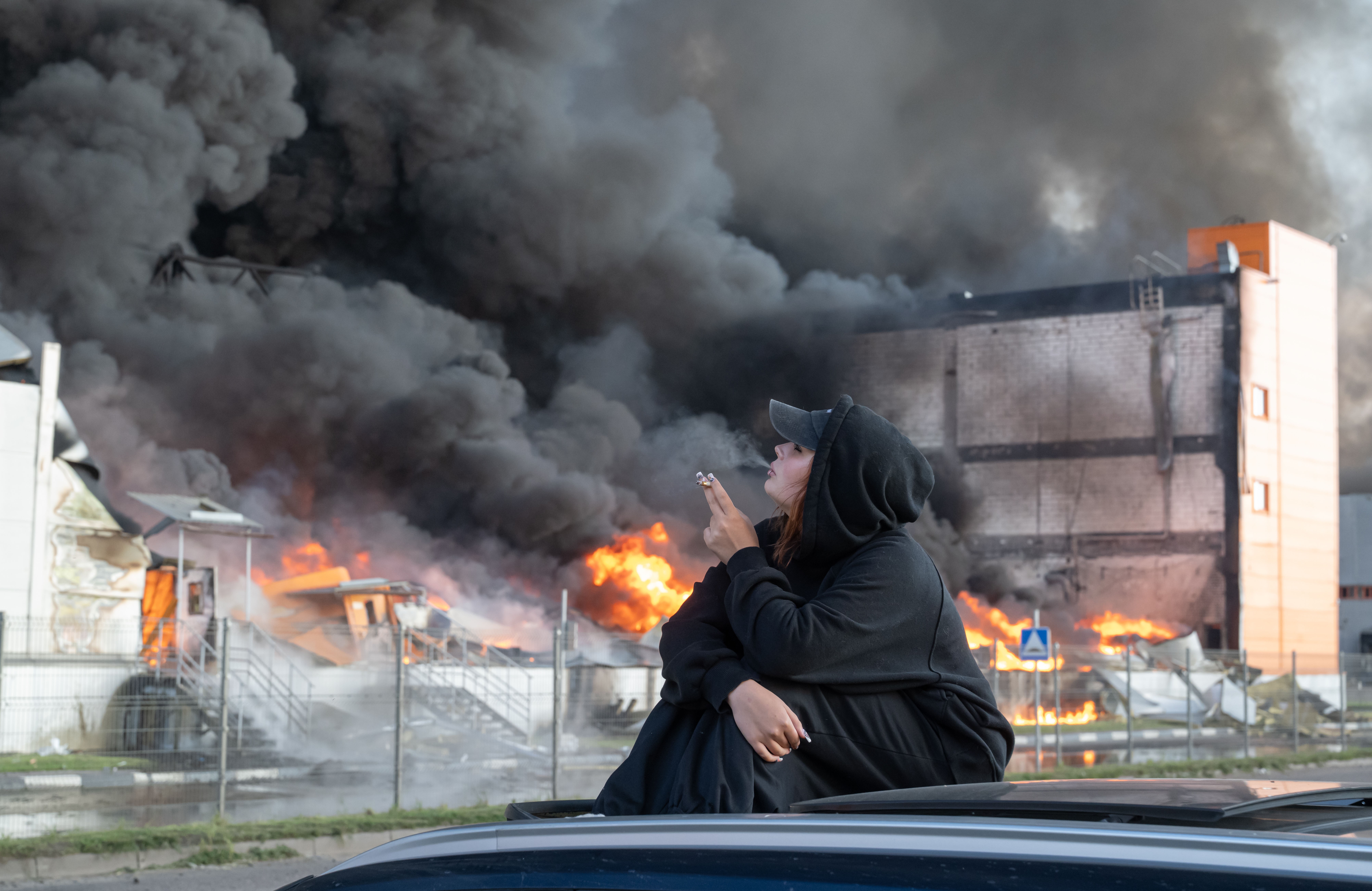 A woman smokes while watching a burning warehouse.