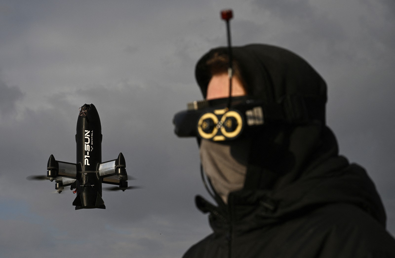 A drone operator, wearing a mask and visor, pilots a drone which hovers in the background.