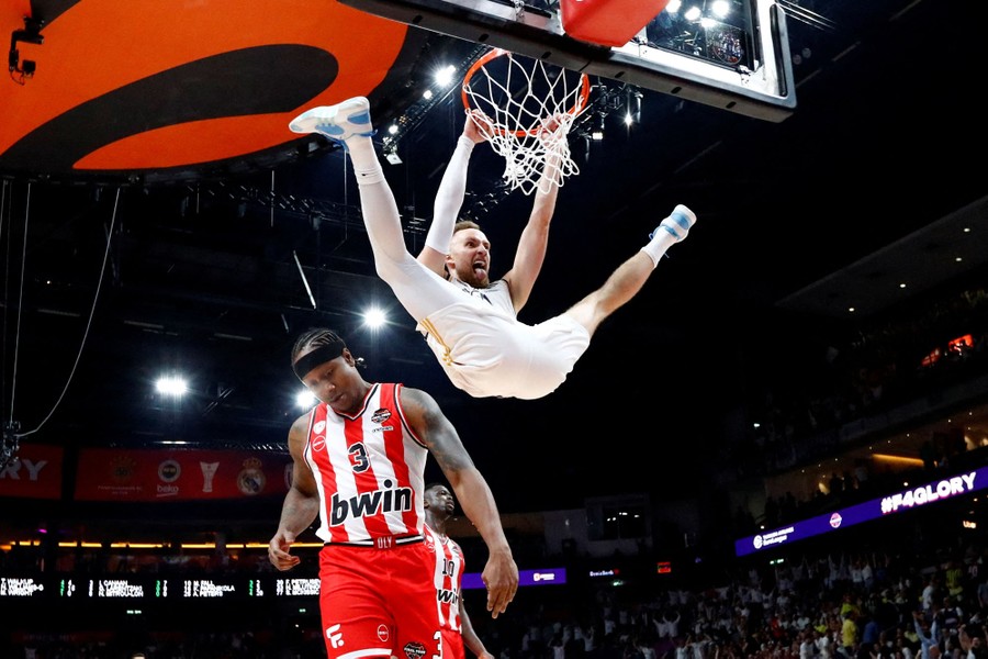 A basketball player makes a face while hanging from a basket and kicking both legs up, above two opponents.