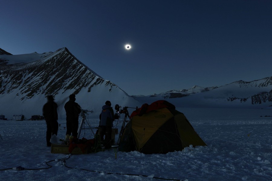People stand in a snowy field with telescopes, beside a tent, under a dark sky, looking up at a total eclipse.