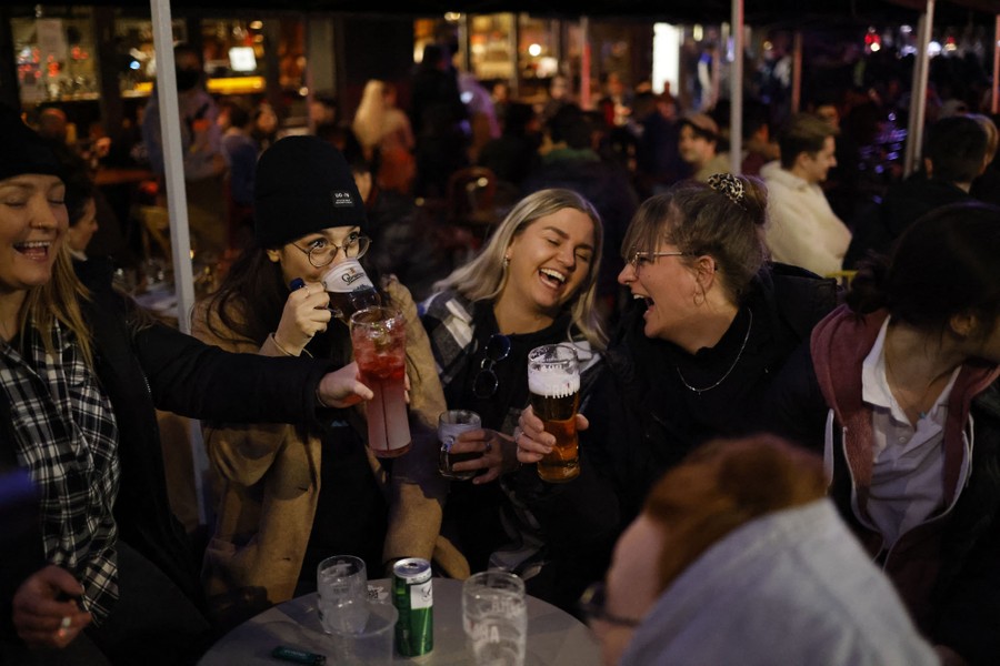 People laugh while drinking, seated at outside tables at a bar.