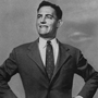 A black-and-white photo of a smiling man in a suit and tie with his hands at his hips