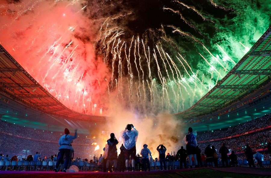 Fireworks erupt around the top of an open stadium as people inside watch.