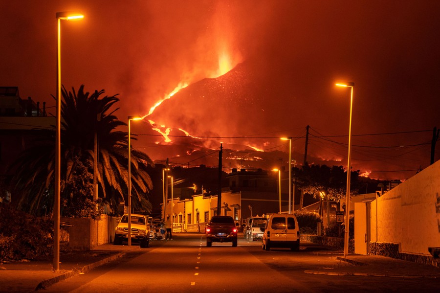 Nighttime view of a city street with lava flowing down a hill in the distance.
