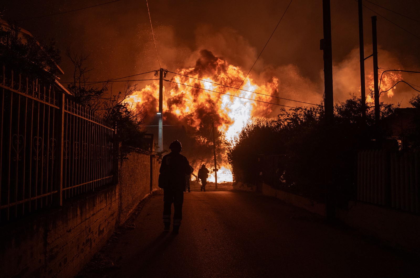 Several firefighters approach a wildfire, walking on a narrow residential road.