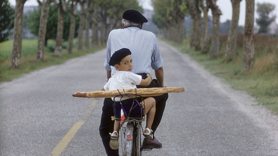 An old man and a young boy, both in berets, ride a bicycle with two baguettes strapped to the back down a tree-lined street; the young boy is looking back over his shoulder at the camera.