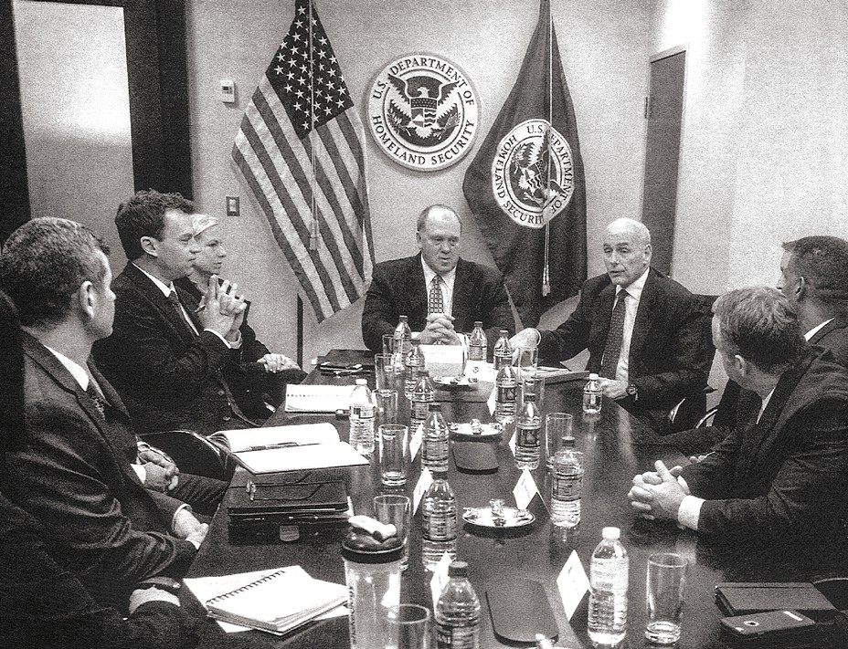 fotografía de una gran mesa de juntas con botellas de agua y papeles rodeada de personal vestido formalmente con banderas de Estados Unidos y del Departamento de Seguridad Nacional (DHS) y un sello en la pared de atrás