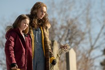 A woman holds flowers and stands with her child in a graveyard.