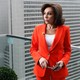 Jeanine Pirro wearing an orange suit and standing on a balcony with a metal and glass railing in front of a cityscape
