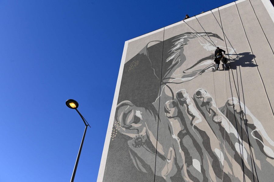 An artist paints a large mural of a person's profile on a building wall.