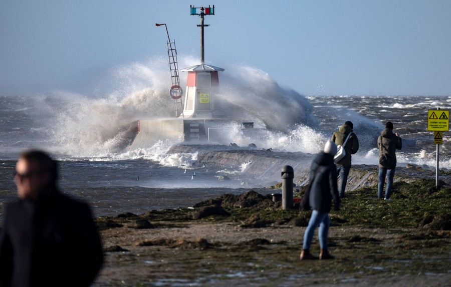 Several people watch as high waves splash over structures in a harbor.