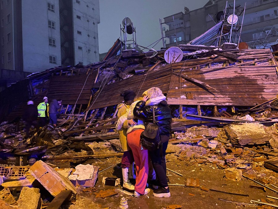 Several people embrace one another, standing in front of the debris of a collapsed building.