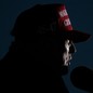 Donald Trump in shadowy profile speaking at a microphone and wearing a red "Make America great again" hat