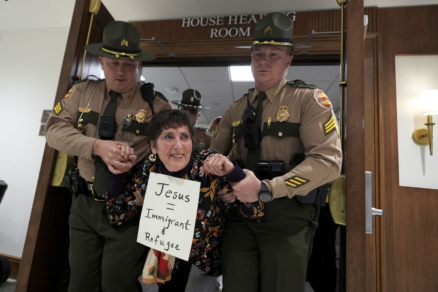Two state troopers escort a woman from a government hearing room. She wears a sign around her neck that reads "Jesus = Immigrant & Refugee."