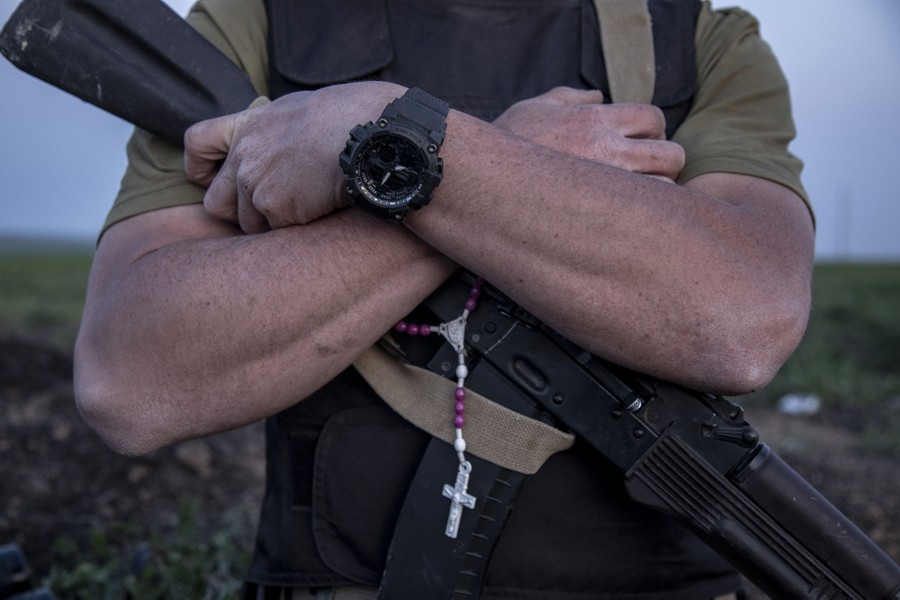 A close view of a soldier wearing short sleeves, with his arms crossed over his rifle and a rosary hanging down from the strap.
