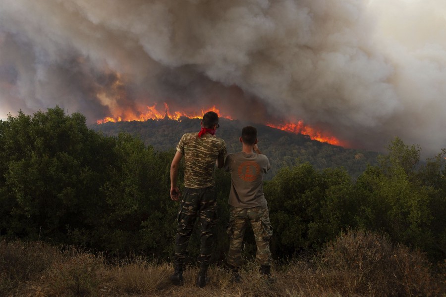 Two people stand on a ridge looking toward a wildfire burning on a nearby hill.
