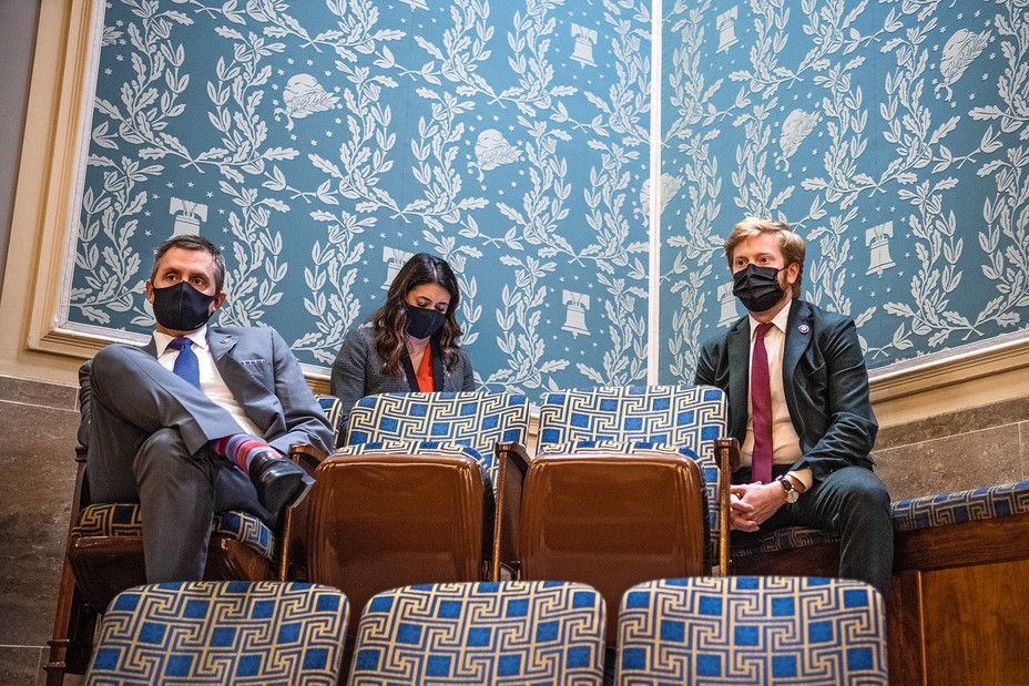 photo of two men and woman in face masks and business attire sitting in the top row of folding seats with blue and white olive-branch and liberty-bell wallpaper behind them