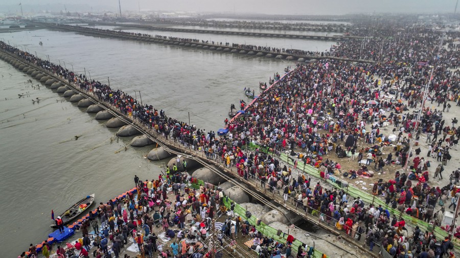 A large crowd gathers on a river shoreline, while some cross several long pontoon bridges.